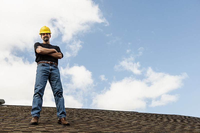 This is a horizontal, color photograph of a male construction worker standing on top of a shingled room. White clouds and blue sky behind him provide copy space.